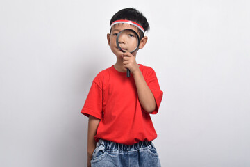 Smart Asian boy holding magnifying glass to search information, isolated on white background. Indonesian Independence Day concept.
