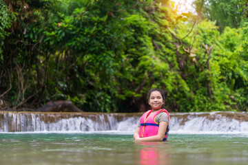 Fototapeta premium Asian child standing in waterfall or kid girl happy smile and wearing life jacket on green nature stream water in forest or wild garden with trees on summer holiday travel at huai mae khamin waterfall
