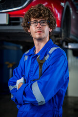 Professional Auto Mechanic in Blue Overalls Holding Wrench in Modern Garage with Vehicle Lifted for Maintenance