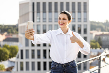 Smiling woman on rooftop using smartphone for selfie with thumbs up. Urban background with buildings and greenery. Casual attire, happy and confident expression.