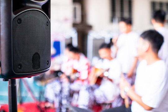 Close-up of a black stage speaker on a stand with a youth acoustic band performing in a blurred background.