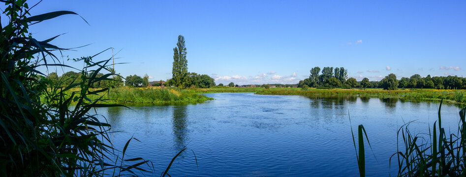 Panoramic photo of a flowing wide river "De Niers" in Germany surrounded by green grass and green trees, with a blue sky with some white clouds.