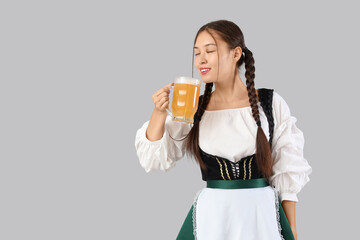 Beautiful young happy Asian Octoberfest waitress with mug of beer on grey background