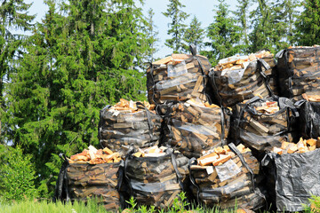 Stacks of cut and split firewood stored in large heavy-duty mesh bags with coniferous forest in the background.