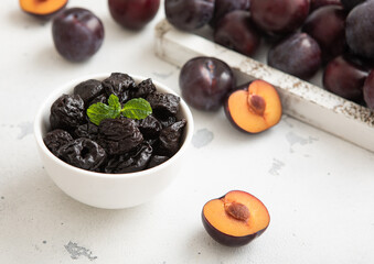 Sweet prunes in glass bowl with ripe plums in wooden box on light kitchen background.