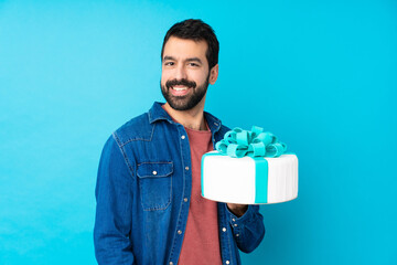 Young handsome man with a big cake over isolated blue background laughing