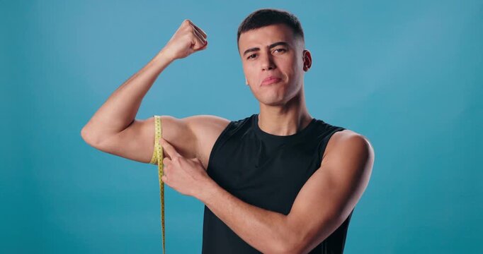 Measuring tape, face and man with muscles in studio for fitness, workout or body building progress. Portrait, strength and male person flex arm biceps for exercise goals or target by blue background.