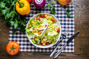 Caesar Salad with cabbage, tomato, cherry tomato, roasted chicken, onion, capsicum, coriander, knife and fork served in dish isolated on napkin top view of italian healthy food on wooden table