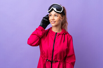Skier teenager girl with snowboarding glasses over isolated purple background keeping a conversation with the mobile phone