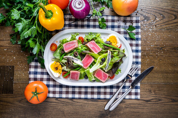 Nicoise Salad with cherry tomato, fish, eggs, olives, herbs, tomato, onion, capsicum, coriander, knife and fork served in dish isolated on napkin top view of italian healthy food on wooden table