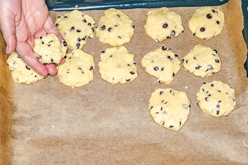 One hand making homemade cookie dough balls with chocolate chips on a baking tray covered with baking paper. Overhead personal point of view.