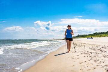 Playing sports on beach in summer. Portrait of young modern woman with long blonde hair in blue top and black shorts and headphones, sunglasses practicing Nordic walking on sandy beach on sunny day. 