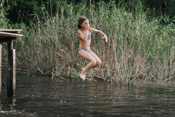 Athletic beautiful teenage girl, child jumping from a wooden bridge, pier into the water outdoors in nature. Photography, portrait, childhood concept, sport.