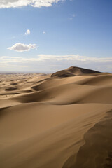 A vast, serene desert landscape with rolling sand dunes in Gobi Gurvan Saikhan National Park under a clear blue sky with scattered clouds.