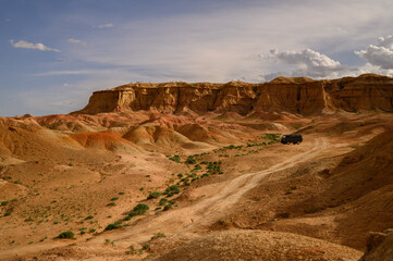A rugged and colorful landscape of the Gobi desert with rocky formations and a vehicle traversing the arid terrain under a partly cloudy sky. Flaming Cliffs – Bayanzag