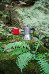 Coffee pot and metal cup on big stone against backdrop of mountain river. Coffee pot on stone plate over blurred nature background.