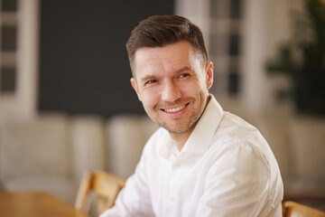 Obraz premium A man in a white shirt smiles warmly, seated indoors with soft, blurred background lighting.
