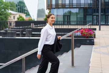 A young woman in business attire walks up stairs in a modern urban setting, smiling confidently.