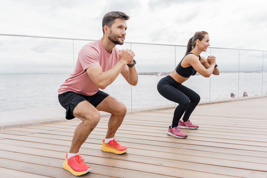 A man and woman perform squats together on a wooden deck by the water, focusing on their workout. - Powered by Adobe