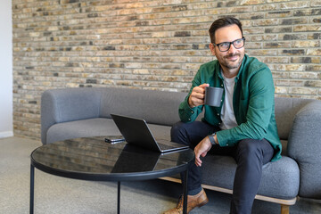 Thoughtful male freelancer enjoying coffee and looking away while relaxing on sofa in modern office