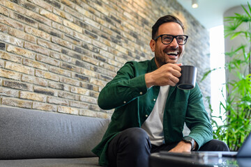 Low angle view of cheerful businessman having coffee and looking away while relaxing on sofa in...