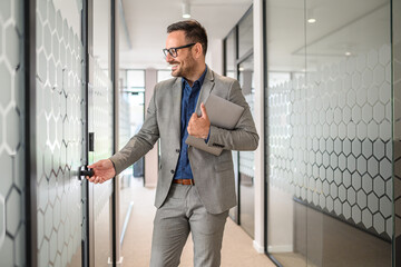 Handsome smiling entrepreneur holding laptop and opening glass door of board room in modern office