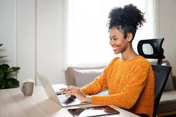 Smiling young African American woman sitting at desk working on laptop, happy millennial female studying online, watching webinar using computer