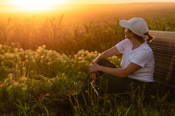 Woman wearing white cap sits next to solar panel at sunset. Wind turbines in the background.