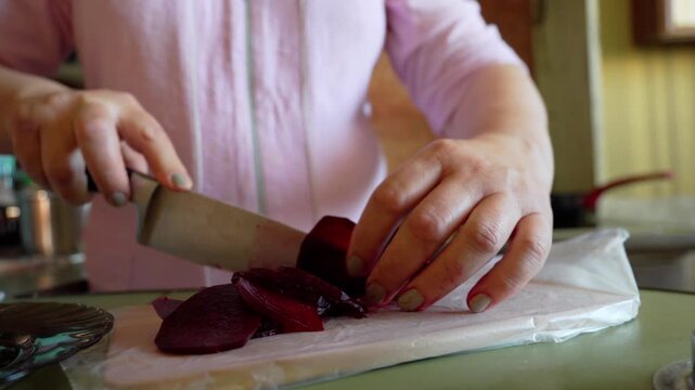 Holds a knife and slices a beetroot on a wooden board. The beetroot is wrapped in plastic wrap.