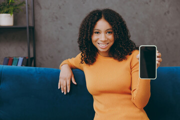 Little kid teen girl wear casual clothes hold use blank screen mobile cell phone sits on blue sofa couch stay at home hotel flat rest relax spend free spare time in living room indoor. Lounge concept.