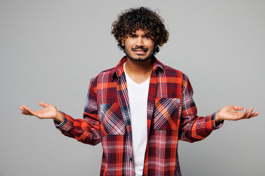 Young sad mad Indian man he wears red shirt casual clothes shrugging shoulders looking puzzled, have no idea, spread hands isolated on plain grey color background studio portrait. Lifestyle concept.