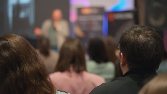 Close-up short of backs of people's heads at conference. Men and women looking at lecturer. Tech-savvy youth absorb new technological trends from speaker at innovation seminar, presenter blurred.