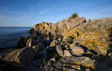 Rocky coastal landscape with large boulders and sunlit cliffs overlooking a calm sea under a clear blue sky on the Danish Island of Bornholm - Powered by Adobe