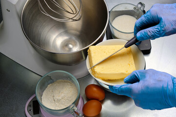 Chef's hands with knife cutting off piece of butter against background of pie ingredients and mixer bowl.