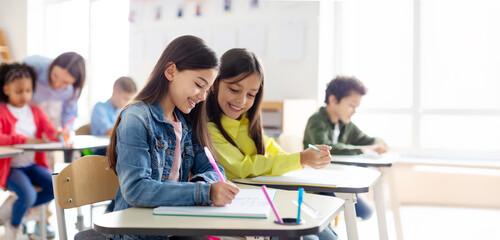 School children sit at desks, writing in copybooks, one girl glances at her friend's test and smiling, studying at school © Home-stock