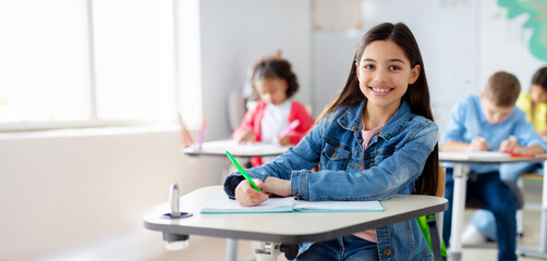 Smiling junior school girl sitting at desk in classroom, writing in notebook, posing and looking at...