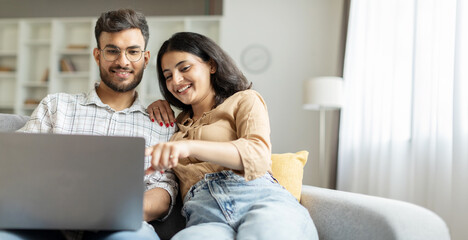 Happy indian couple using laptop, surfing web, woman pointing at screen, sitting on sofa. 