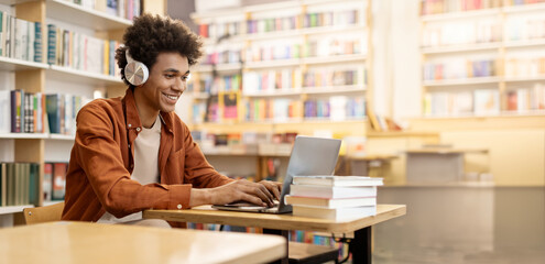 Dedicated smiling black male student engaged in online learning at college library, using laptop to study, surrounded by bookshelves