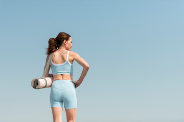 Fit, sporty woman carrying an exercise mat, outdoors, rear view.