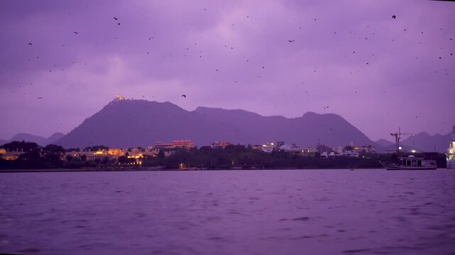 Moving boat shot over water of lake pichola showing leela palace oberoi with Sajjan Garh monsoon place in the distance at dusk with the amazing tourist landmarks in Udaipur