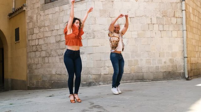 Two joyful dancers perform an energetic salsa routine on a sunlit urban street against a classic stone wall backdrop