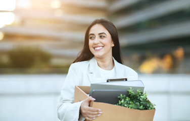 A young woman in a white blazer smiles as she carries a box containing her belongings. She is leaving her office building and heading towards a new adventure, copy space