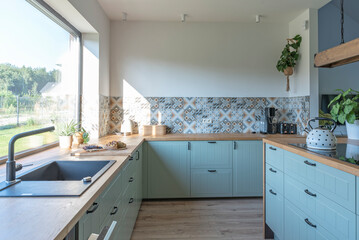 Modern kitchen interior with kitchen sink near window, stylish furniture and pattern tile on the wall. Home design in new house.