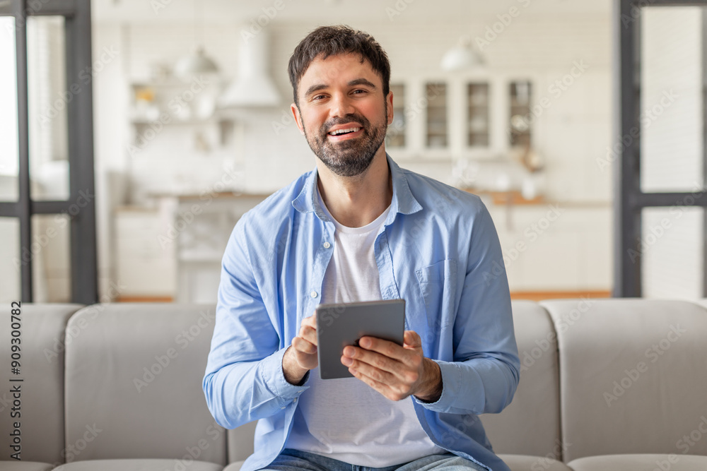Poster a man sits on a gray couch in his home and smiles while holding a tablet computer in his hands, webs