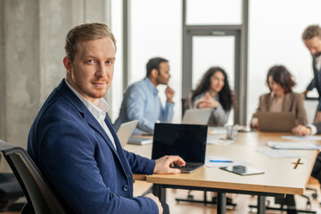 A young businessman in a blue suit is working at a desk in a modern office. He is looking directly at the camera with a confident expression. He is sitting at a desk with a laptop computer