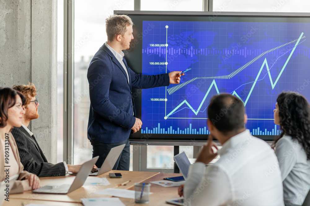 Fototapete A businessman in a suit points to an upward trending line graph on a large screen while presenting to his team during a meeting. His team sits around a table with laptops open