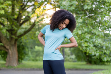 Young African American athlete holding lower back showing pain while exercising outdoors. Woman wearing blue t-shirt and black leggings standing among trees in park.