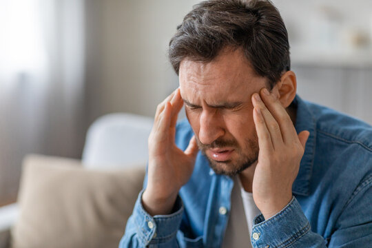 A man in a blue shirt is sitting on a couch. He is holding his head in his hands, and his face is contorted in pain. He appears to be suffering from a headache, closeup, copy space
