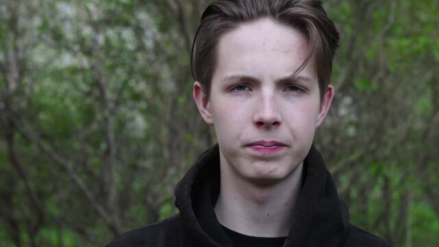 Close-up of a young boy's face against a background of branches. Cute teenage boy in a black hooded biker spitting sideways.High quality FullHD footage