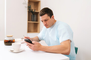 Male patient in medical coat resting, eating, using smartphone before long operation in clinic. Caucasian young man enjoy eating food sitting at table in hospital room with light interior.